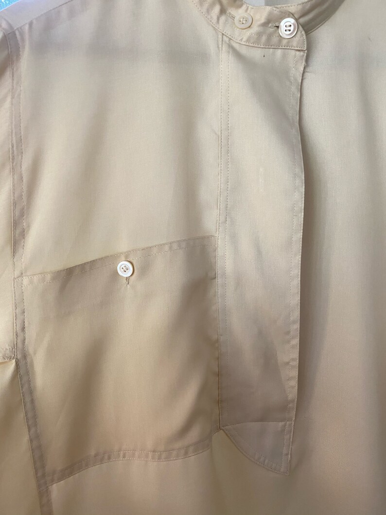 May include: A close-up of a beige button-down shirt with a single patch pocket on the left side. The shirt has a mandarin collar and a single button closure at the neck.