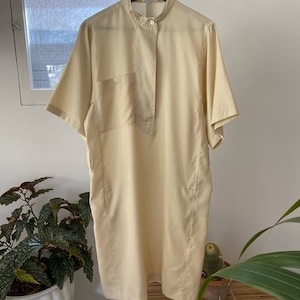 May include: A pale yellow short-sleeved collared dress with a single button closure at the neck and a patch pocket on the left side.