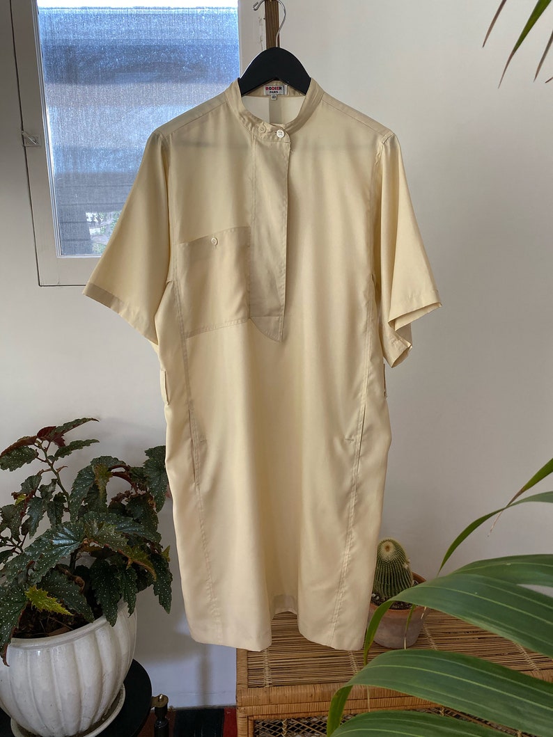 May include: A pale yellow short-sleeved collared dress with a single button closure at the neck and a patch pocket on the left side.