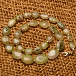 May include: A beaded necklace featuring oval-shaped, light green and white stones. The stones are strung together with small silver-colored beads and a silver clasp. The necklace is displayed on a brown, textured surface.