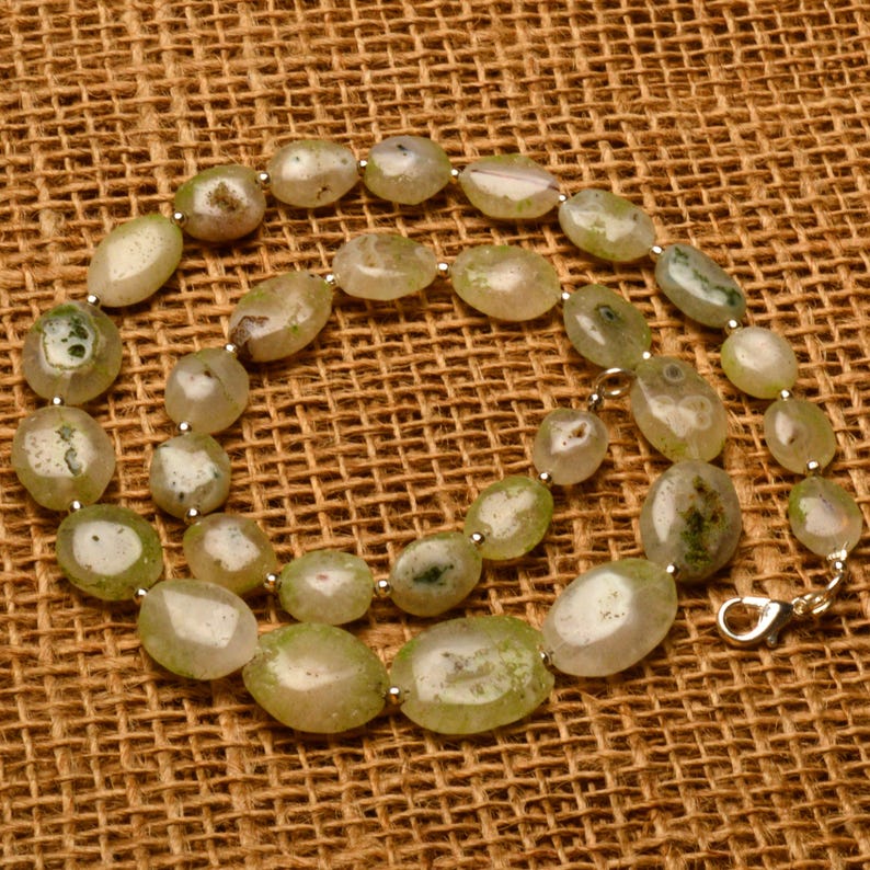 May include: A beaded necklace featuring oval-shaped, light green and white stones. The stones are strung together with small silver-colored beads and a silver clasp. The necklace is displayed on a brown, textured surface.