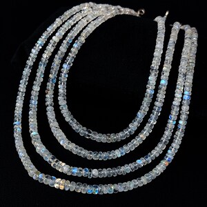 Puede incluir: Cinco hilos de collares de piedras preciosas claras y facetadas con un sutil brillo iridiscente. Las cuentas est&aacute;n ensartadas, creando un efecto en capas. El fondo es negro, resaltando los collares.