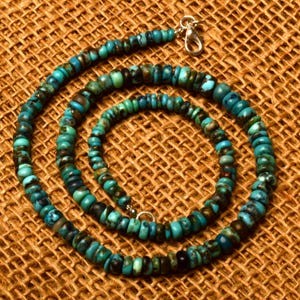 May include: A turquoise beaded necklace with a silver clasp. The necklace features small, round beads in shades of turquoise, blue, and brown. The necklace is displayed on a textured, brown surface.