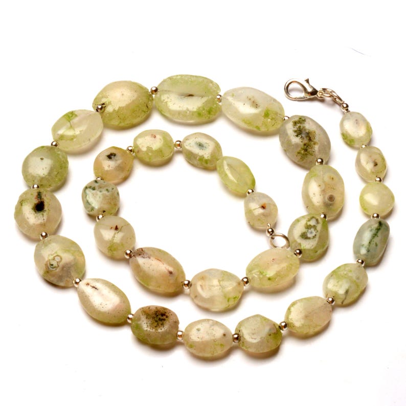 May include: A necklace featuring light green and cream-colored oval-shaped beads. The beads have a speckled appearance and are strung together with small gold-colored beads. The necklace is secured with a silver-colored clasp.
