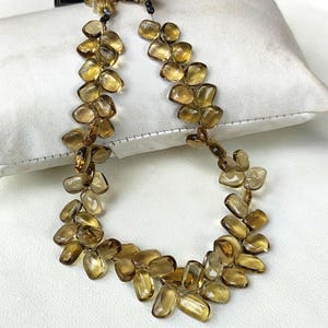 May include: A necklace featuring a series of amber-colored, irregularly shaped stones. The stones are strung together, creating a flowing, organic design. The necklace is displayed on a white surface, highlighting the warm tones of the gemstones.