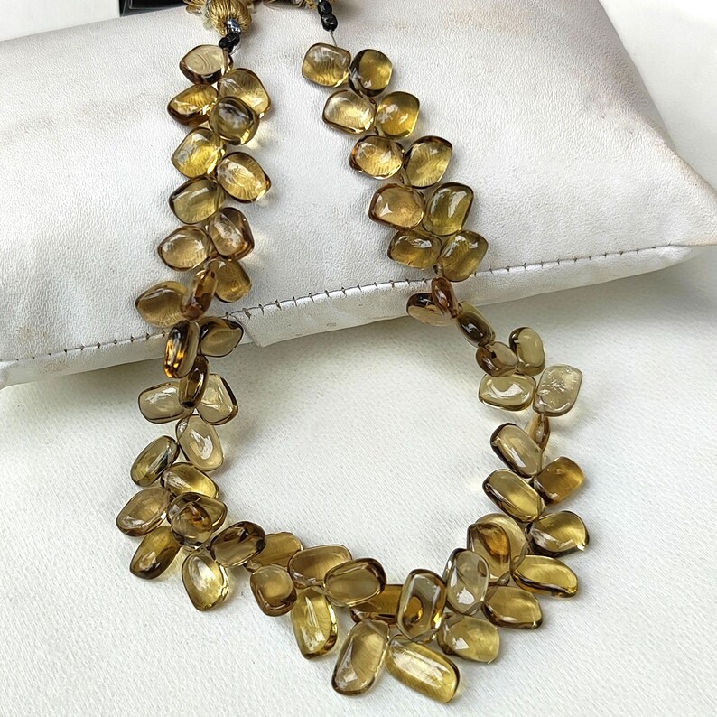 May include: A necklace featuring a series of amber-colored, irregularly shaped stones. The stones are strung together, creating a flowing, organic design. The necklace is displayed on a white surface, highlighting the warm tones of the gemstones.