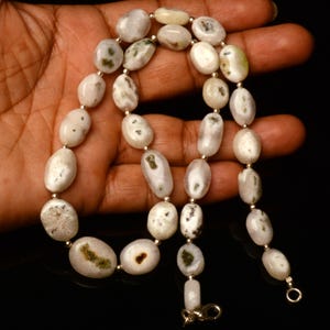 May include: A necklace featuring oval-shaped, off-white beads with green and brown inclusions. The beads are strung together with small silver-colored beads and a silver-colored clasp. The necklace is displayed against a dark background.