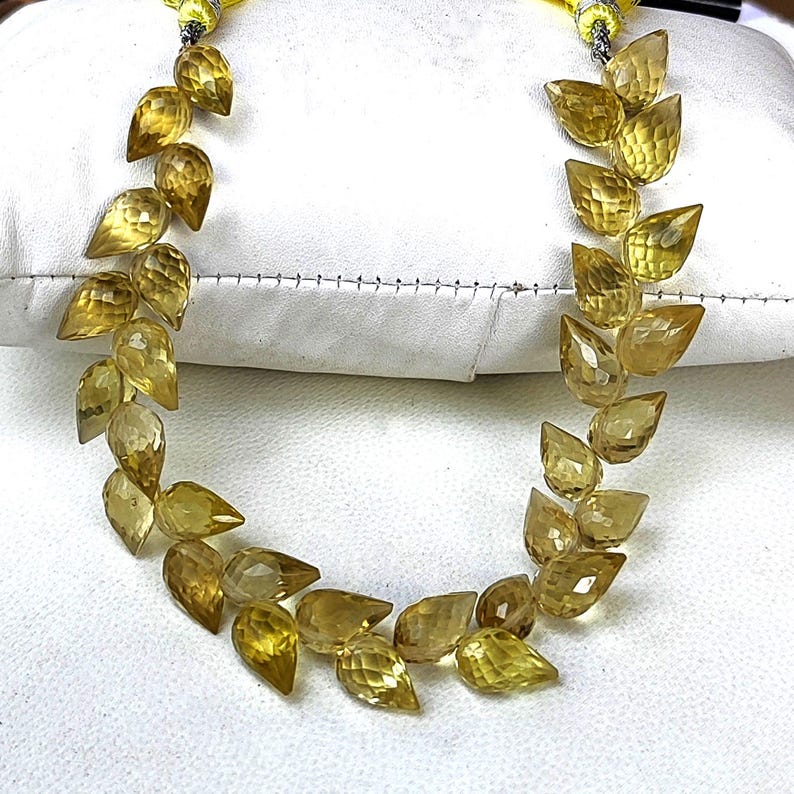 May include: A strand of yellow gemstone beads in a teardrop shape. The beads are strung on a cord and are arranged in a pattern of alternating directions.
