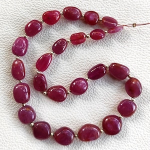 May include: A strand of polished, oval-shaped ruby beads. The beads are a deep, rich red color, strung together with small silver-colored spacers. The beads are smooth and have a natural, organic shape, suggesting they are genuine gemstones.