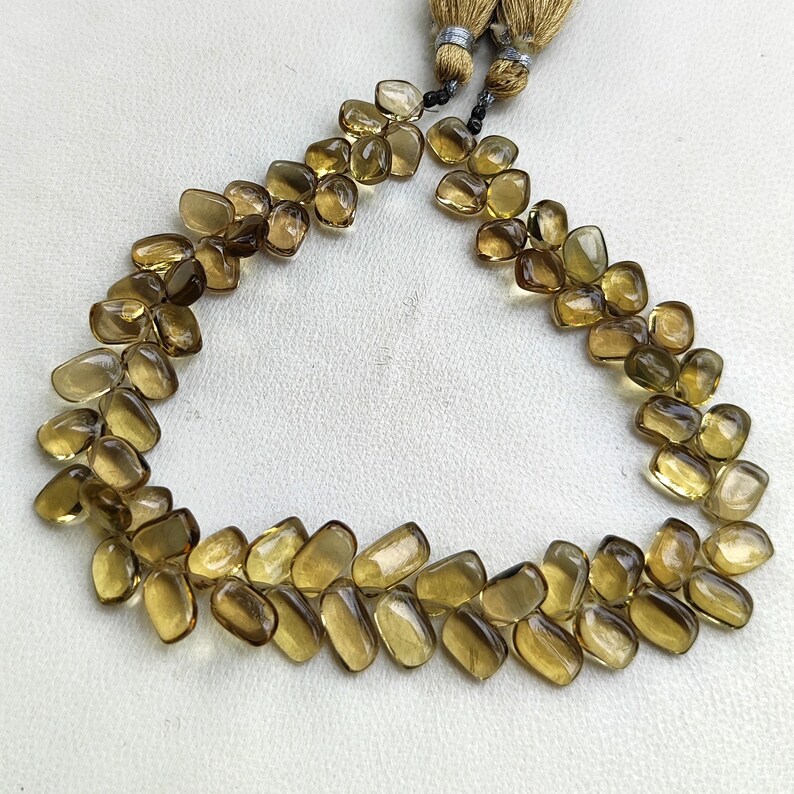 May include: A necklace made of polished, amber-colored gemstones. The stones are irregularly shaped and strung together, forming a curved design. The necklace has a tassel at each end, made of tan-colored threads.