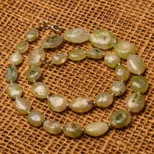 May include: A necklace featuring irregularly shaped, light green and white beads. The beads are strung together with silver-colored metal links and a clasp. The necklace is displayed on a brown, textured surface.