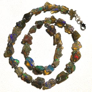 Pode incluir: Um colar com pedras de opala brutas, não cortadas, em tons de castanho, verde e azul. As pedras são enfiadas com pequenas contas de prata e um fecho de prata. As pedras de opala têm uma textura natural e áspera.