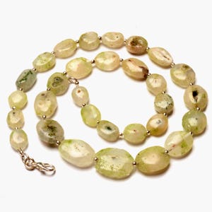 May include: A necklace featuring irregularly shaped, light green and cream-colored beads. The beads are strung together with small silver-colored connectors and a clasp. The necklace has a natural, organic appearance.