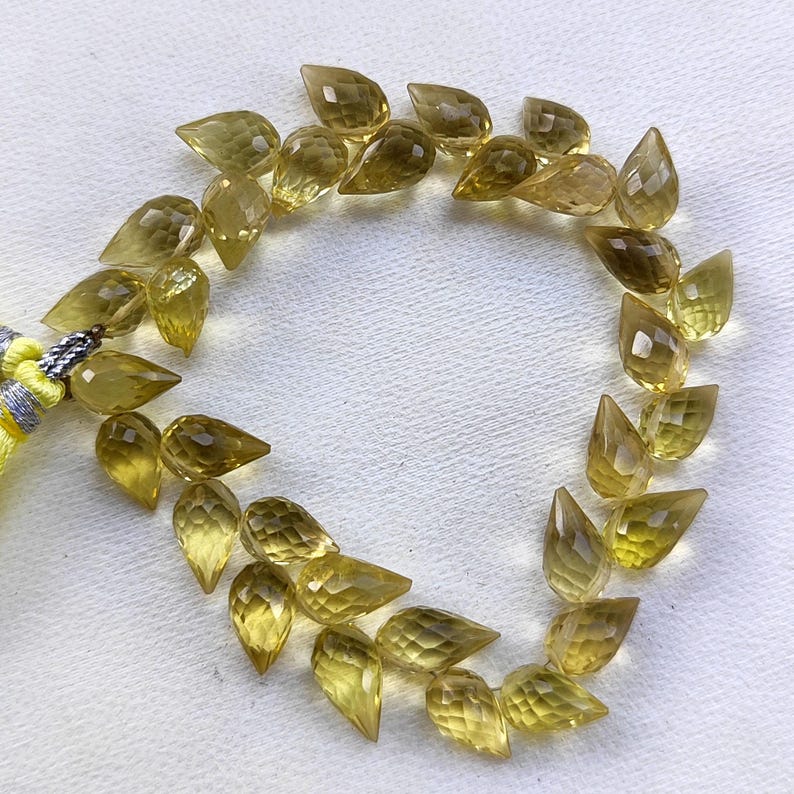 May include: A strand of faceted teardrop-shaped beads in a light yellow color. The beads are arranged in a circle on a white surface.