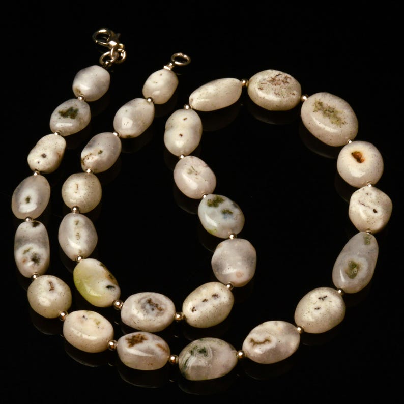 May include: A necklace featuring oval-shaped, off-white beads with subtle green and brown markings. The beads are strung together with small gold-colored spacers and a gold-colored clasp. The necklace is displayed against a black background.