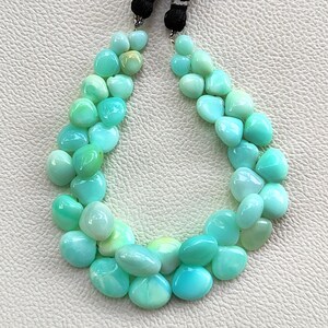 May include: A strand of polished, teardrop-shaped mint green gemstones arranged in a U-shape. The smooth, opaque stones feature subtle variations in light green and turquoise hues, secured at the top by black cord ends with silver-toned metal accents.