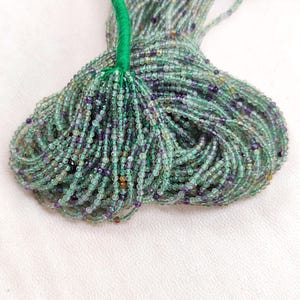 May include: A strand of green, purple, and clear round gemstone beads. The beads are small and uniform in size. The strand is tied together with a green string.