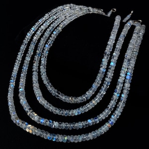 Puede incluir: Cinco hilos de collares de piedras preciosas claras e iridiscentes. Las cuentas son peque&ntilde;as y redondas, con destellos de azul y blanco. Los collares se exhiben sobre un fondo negro, mostrando los delicados detalles de la joyer&iacute;a.