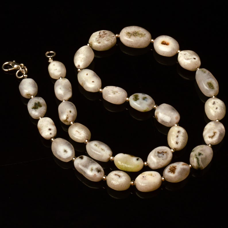 May include: A necklace featuring large, oval-shaped beads in shades of cream, beige, and gray, with subtle green and brown inclusions. The beads are strung together with small gold-colored spacers and a gold-toned clasp. The necklace is displayed against a black background.