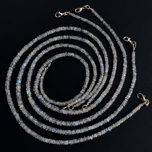 Puede incluir: M&uacute;ltiples hilos de delicados collares de cuentas de color claro con cierres plateados. Las cuentas tienen una sutil iridiscencia, con toques de azul y blanco. Los collares est&aacute;n dispuestos en un patr&oacute;n en capas y fluido sobre un fondo negro.