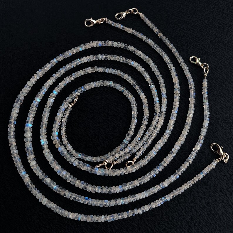 Puede incluir: M&uacute;ltiples hilos de delicados collares de cuentas de color claro con cierres plateados. Las cuentas tienen una sutil iridiscencia, con toques de azul y blanco. Los collares est&aacute;n dispuestos en un patr&oacute;n en capas y fluido sobre un fondo negro.