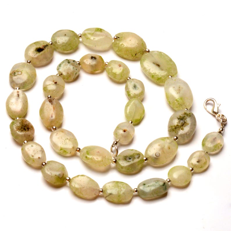 May include: A necklace featuring oval-shaped, light green gemstones with silver-colored beads and a clasp. The stones have a natural, organic appearance with variations in color and texture, creating a unique jewelry piece.