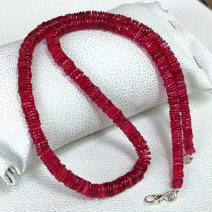May include: A necklace composed of numerous small, flat, circular, deep red beads strung together. The strand features a silver-toned metal lobster claw clasp at the end, providing a secure closure for the piece of jewelry.