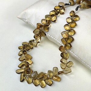 May include: A necklace composed of numerous, irregularly shaped, amber-colored gemstones. The stones are strung together, forming a flowing, organic design. The necklace rests on a white surface, with gold-colored tassels visible at the top.