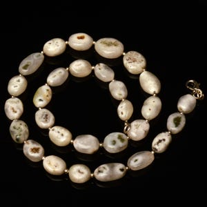 May include: A necklace featuring oval-shaped, off-white beads with subtle green and brown inclusions. The beads are strung together with small gold-colored spacers and a gold-tone clasp. The necklace is displayed against a black background.