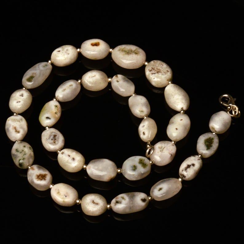 May include: A necklace featuring oval-shaped, off-white beads with subtle green and brown inclusions. The beads are strung together with small gold-colored spacers and a gold-tone clasp. The necklace is displayed against a black background.
