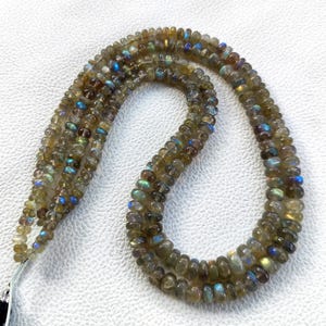 Puede incluir: Cadenas de cuentas de piedra preciosa labradorita. Las cuentas son una mezcla de marrón, gris y verde con destellos de azul y dorado. Las cuentas son pequeñas y redondas, ensartadas para formar un collar.