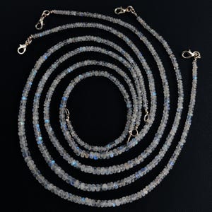 Puede incluir: M&uacute;ltiples hilos de delicados collares de cuentas con un brillo iridiscente. Las peque&ntilde;as cuentas redondas muestran tonos blancos y azules. Cada hilo tiene un cierre dorado. Los collares est&aacute;n dispuestos en un patr&oacute;n circular sobre un fondo negro.