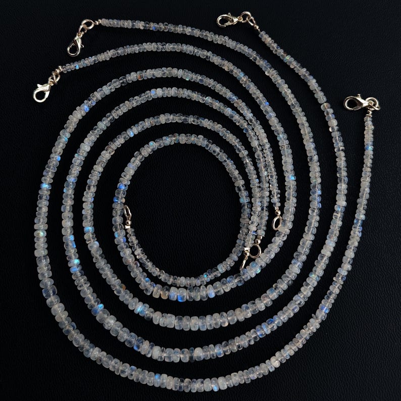 Puede incluir: M&uacute;ltiples hilos de delicados collares de cuentas con un brillo iridiscente. Las peque&ntilde;as cuentas redondas muestran tonos blancos y azules. Cada hilo tiene un cierre dorado. Los collares est&aacute;n dispuestos en un patr&oacute;n circular sobre un fondo negro.