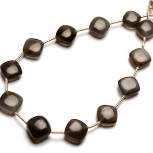 Puede incluir: Una hebra de cuentas de piedras preciosas de color gris oscuro con forma cuadrada. Las cuentas están enhebradas en un alambre dorado.