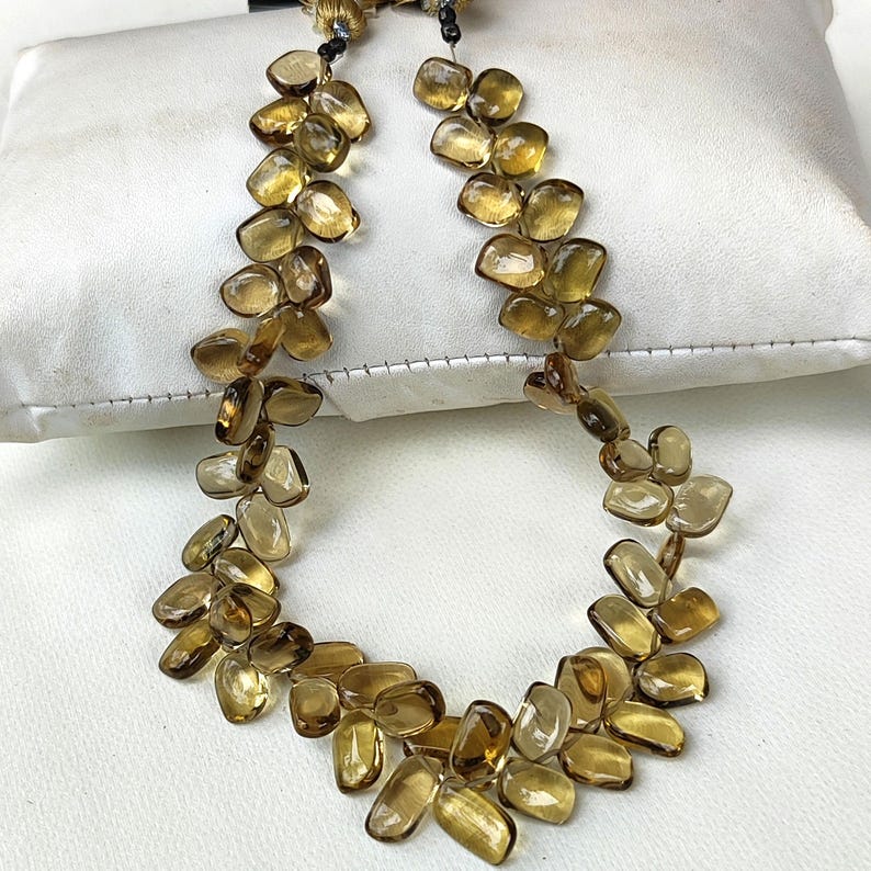 May include: A necklace featuring numerous amber-colored, irregularly shaped stones. The stones are strung together, creating a flowing, organic design. The necklace is displayed on a white surface, highlighting the warm tones of the gemstones.