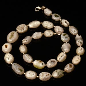 May include: A necklace featuring oval-shaped, off-white stones with subtle green and brown inclusions. The stones are strung together with small gold-colored beads and a gold-tone clasp. The necklace is displayed against a black background.