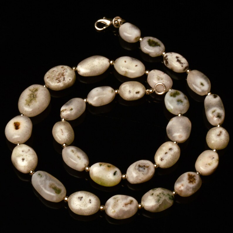 May include: A necklace featuring oval-shaped, off-white stones with subtle green and brown inclusions. The stones are strung together with small gold-colored beads and a gold-tone clasp. The necklace is displayed against a black background.