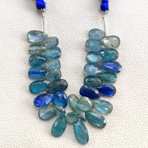 Puede incluir: Dos hilos de cuentas de piedras preciosas azules con una variedad de tonos, desde aguamarina claro hasta índigo intenso. Las cuentas en forma de lágrima están ensartadas en un alambre plateado y unidas a una borla azul.