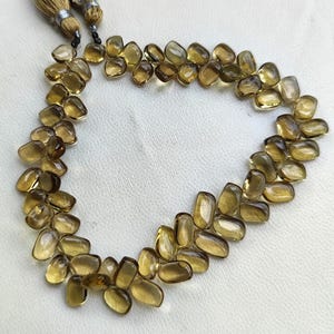 May include: A necklace of polished, amber-colored gemstones. The stones are irregularly shaped and strung together, forming a necklace. The necklace is displayed on a white surface. Tassels hang from the top of the necklace.
