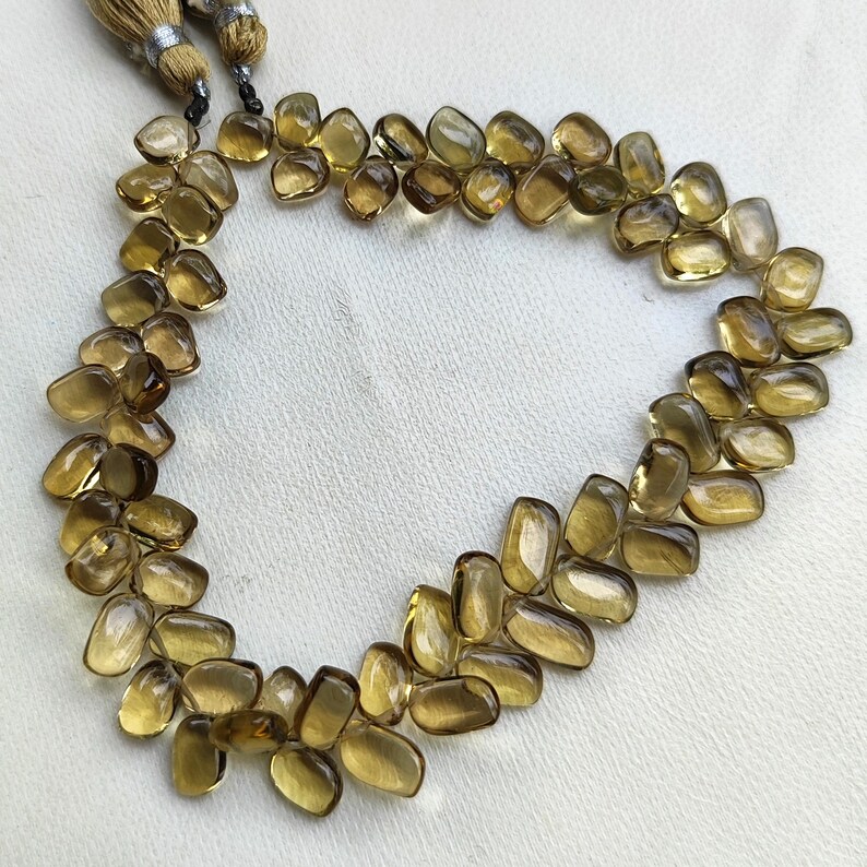 May include: A necklace of polished, amber-colored gemstones. The stones are irregularly shaped and strung together, forming a necklace. The necklace is displayed on a white surface. Tassels hang from the top of the necklace.