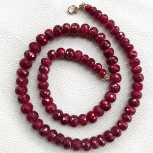 以下が含まれることがあります： ファセット加工された深紅色のルビービーズで構成されたネックレス。ビーズはつながっており、金色の留め金が付いた連続したループを形成しています。ネックレスは白い表面に表示されています。
