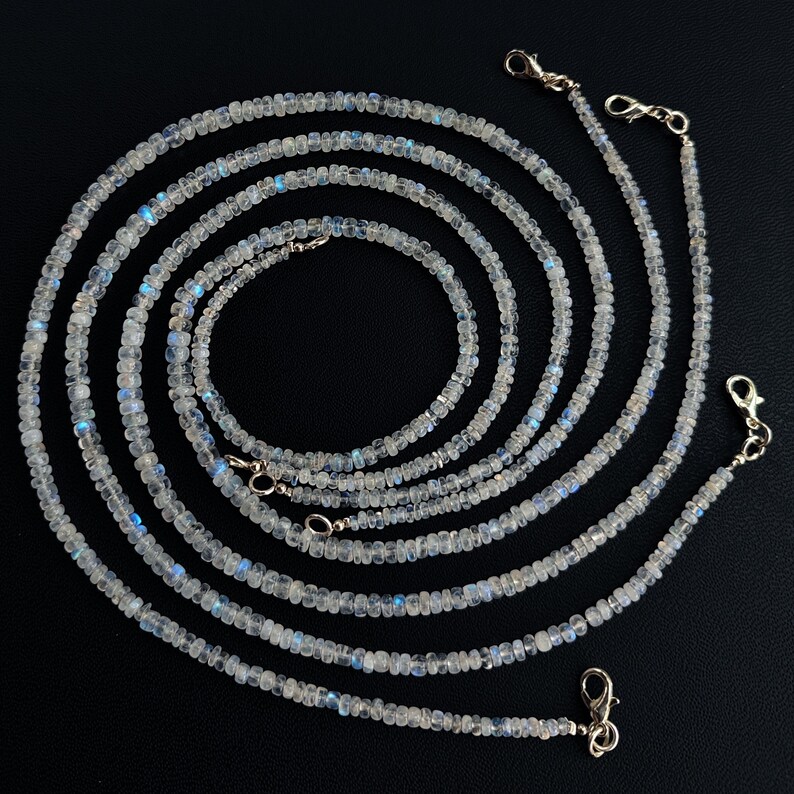 Puede incluir: Varias hebras de delicados collares de piedras preciosas blancas con destellos azules iridiscentes. Cada collar est&aacute; compuesto por peque&ntilde;as cuentas redondas y presenta un cierre plateado. Los collares est&aacute;n dispuestos en un patr&oacute;n en espiral sobre un fondo negro.