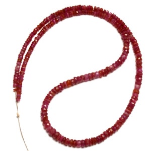 May include: A strand of faceted ruby beads in varying shades of red. The beads are strung together, creating a necklace-like arrangement. The gemstones have a slightly irregular shape and are displayed against a white background.