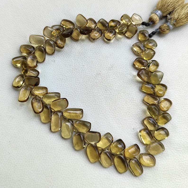 May include: A necklace of polished, light brown citrine gemstones. The stones are irregularly shaped and strung together, forming a curved design. Two gold-colored tassels are at the top of the necklace.
