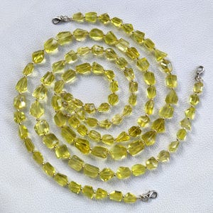 Può includere: Una collana con pietre preziose giallo-verdi irregolari e sfaccettate. La collana è disposta a spirale, con chiusure argentate a ciascuna estremità. Le pietre preziose variano per dimensioni e forma, creando un aspetto unico e organico.
