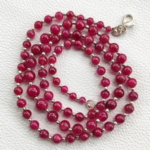 Peut inclure: Un collier à plusieurs rangs composé de perles rondes rouge foncé de différentes tailles. Les perles sont séparées par des entretoises argentées et un fermoir argenté. Le collier est présenté sur une surface blanche.