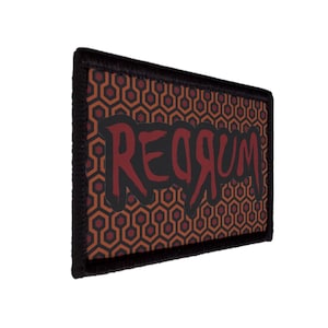 Op de afbeelding: Een rechthoekige patch met een zwarte rand. De patch toont het woord "REDRUM" in een donkerrood, gestileerd lettertype. De achtergrond heeft een herhalend geometrisch patroon in bruin- en oranje tinten.
