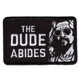 Op de afbeelding: Zwart rechthoekig embleem met de tekst "THE DUDE ABIDES" in witte letters. Een zwart-wit illustratie van een man met een zonnebril en een baard staat aan de rechterkant van het embleem.