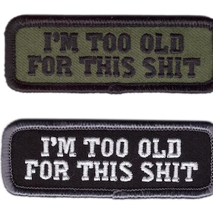 Könnte beinhalten: Zwei rechteckige Aufnäher mit gesticktem Text. Der obere Aufnäher ist olivgrün mit schwarzem Text "I'M TOO OLD FOR THIS SHIT". Der untere Aufnäher ist schwarz mit weißem Text, der dasselbe aussagt.