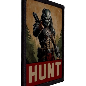 Pode incluir: Remendo retangular com uma ilustração do personagem Predator. A imagem mostra o Predator fazendo um gesto de polegar para cima. A palavra "HUNT" é exibida em letras em negrito sobre um fundo vermelho.